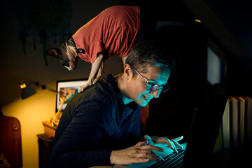Woman with cat climbing on her back while working on laptop at night