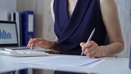 Businesswoman with blue dress is using dark calculator and taking notes in her office. Taxes and...