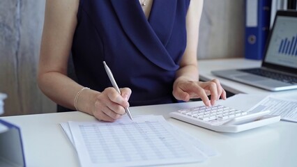 Businesswoman with dark blue dress is using white calculator and taking notes in her office. Taxes...
