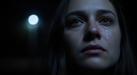 Tearful woman's face in dark, illuminated by a distant light, conveying sadness and vulnerability