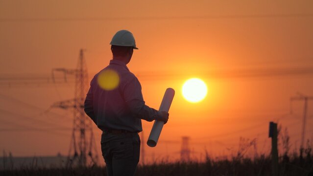 Power engineer working on power transmission line supports repair, sunset. Engineer man working on high voltage power plant construction project. Civil engineer reading drawing. Solar energy, nature
