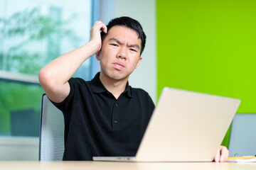 A young man scratches his head in confusion while looking at a laptop screen in a modern classroom. Showing problem solving, online learning frustration, or thinking through difficult tasks.