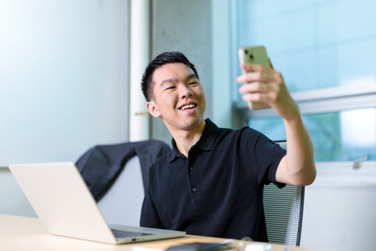 A smiling young man takes a selfie with his smartphone while sitting at a desk with a laptop in a classroom. Relaxation while studying.