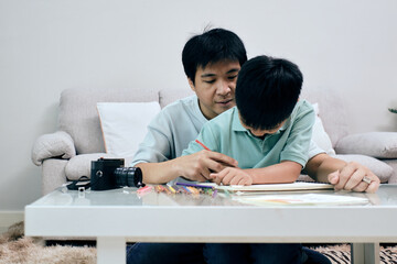 Father sits closely with his young son at a table, guiding him as he works on a drawing with...