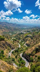 Obraz premium A winding mountain road descends a steep hillside under a vibrant blue sky with fluffy white clouds, showcasing a scenic, natural landscape