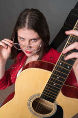 Young female guitarist holding six string classical guitar and looks at camera, lowering her glasses and holding them by temple with one hand. Confidence woman musician dressed in red leather jacket