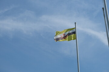 Brunei Darussalam's yellow, black, and white national flag with red crest flying on a tall flagpole under a clear blue sky.
