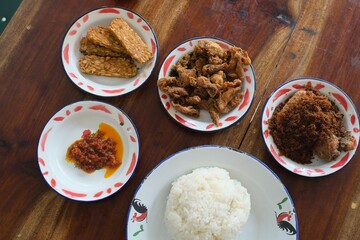 Complete Indonesian meal featuring steamed rice, ayam goreng (fried chicken), tempeh, sambal, and vegetables served on traditional enamel plates.
