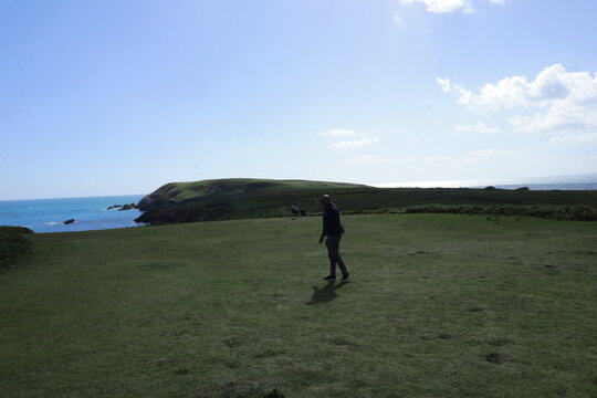 Man walking across wide grassy coastal field with ocean and headland in background under bright sky, expansive travel landscape with copy space, freedom concept and peaceful outdoor lifestyle in remot