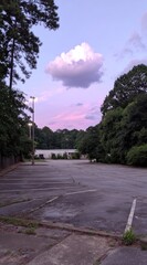 Evening view of an empty, rundown parking lot bordered by green trees with a single white cloud floating in a pink and blue sky at twilight