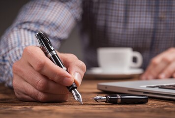 A man's hand writes with a fountain pen on a wooden desk beside a laptop and a cup of coffee, another pen is lying on the desk