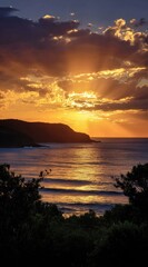 Seascape at sunset. The sky is a fiery orange and purple, with sun rays breaking through clouds. The water reflects the golden light. Hazy mountains in back