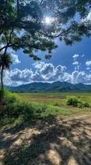 Scenic landscape of verdant fields leading to rolling mountains under a bright sky with fluffy clouds, partially framed by tree branches and foliage