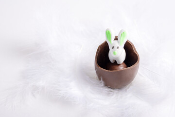 White Easter Bunny in a cracked chocolate egg in a white soft feather nest. Easter marzipan rabbit in a cracked chocolate egg among soft fluffy white feathers. Sweet Easter tradition.