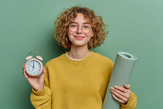 Cheerful young woman with curly hair and glasses holds an alarm clock and yoga mat against a green background. Concept of healthy lifestyle, workout routine and time management