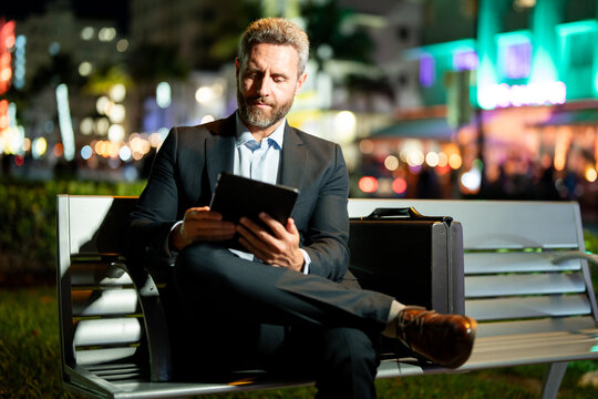 Online business. Businessman working on tablet outdoors. Businessman with tablet outdoor. Businessman with tablet on night street. Businessman using tablet, sit on bench. Night business. Remote work.