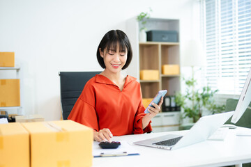 Young Asian woman using calculator and laptop in modern office with boxes. Ideal for themes like...