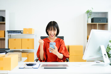 Smiling Asian woman raises fist in joy while holding smartphone. Perfect for business success,...