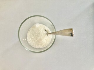 top view shot of a clear glass cup of white sugar and a silver spoon put on a white table