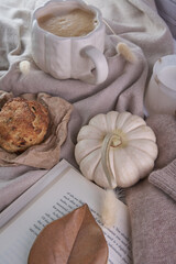 Warm fall lifestyle composition—pumpkin‑shaped white mug of coffee, white mini pumpkin, soft knit sweater and frosted candle arranged on layered neutral fabrics with a rustic pastry, and dried leaves 