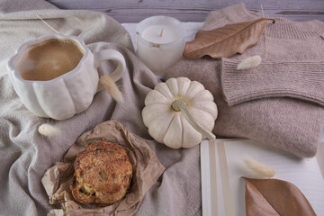 Warm fall lifestyle composition—pumpkin‑shaped white mug of coffee, white mini pumpkin, soft knit sweater and frosted candle arranged on layered neutral fabrics with a rustic pastry, and dried leaves 