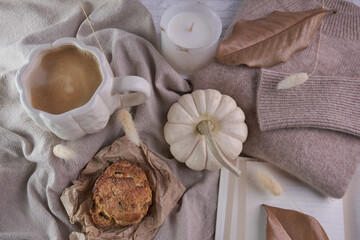 Warm fall lifestyle composition—pumpkin‑shaped white mug of coffee, white mini pumpkin, soft knit sweater and frosted candle arranged on layered neutral fabrics with a rustic pastry, and dried leaves 