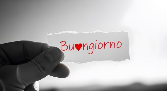 Hand holding a torn piece of paper with the word buongiorno and a heart symbol written in red ink, against a bright, hazy sky