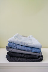 Minimalist product photo of neatly folded light, medium, and dark wash jeans stacked on a white dresser against a soft green wall, modern fashion flat lay and wardrobe organization concept