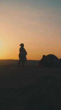 silhouette of overweight woman dancing near camping friends at bonfire on river bank against setting sun low angle shot