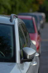 A silver car is in focus in the foreground, showcasing its side mirror and surface reflection. A line of various parked cars is blurred in the background.