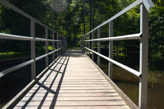 Close-up of a pedestrian bridge. The structure is located above the water on a green background. Gray metal railings and a wooden deck, which is illuminated by sunlight.