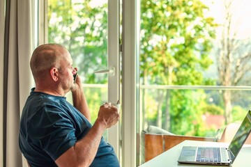 Middle-aged man with laptop, drinking coffee, talking on phone, enjoying window view.
