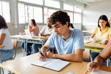 Portrait of young student boy writing on notebook sitting at desk in classroom. Education and back to school concept