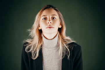 A young woman in a turtleneck sweater and blazer poses against a dark green background. Confident...