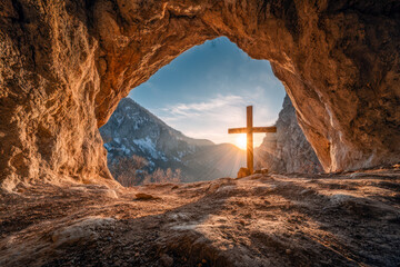 Wooden cross silhouetted by the rising sun seen from inside a rocky cave opening with mountain landscape in the background during early morning light