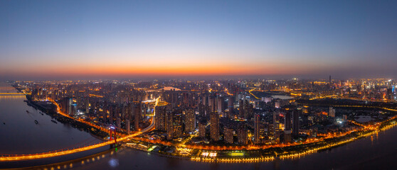 Wuhan, China cityscape at twilight with glowing city lights illuminating the urban landscape and a river reflecting the sky's warm hues.