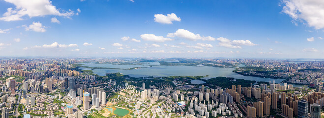A vast city unfolds with many buildings next to a large blue lake under a bright sky with white clouds, capturing a wide urban landscape.