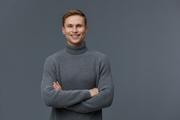 Smiling young man wearing gray turtleneck sweater posing confidently on plain gray background...