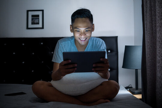Asian man cheerfully enjoying leisure time in a cozy setting playing mobile gaming on his gadget.