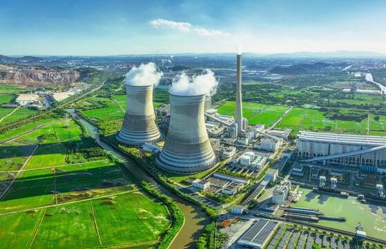 Aerial view of a large thermal power station with cooling towers emitting steam, situated in a green rural landscape with farmland.