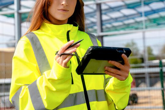 Construction site worker uses tablet for project management in bright safety gear during daylight hours - Powered by Adobe