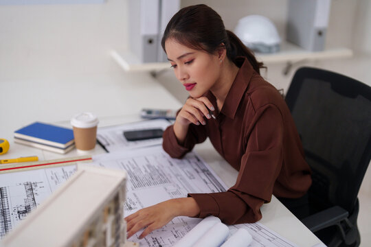 Asian woman architect reviewing building blueprints architectural plans in modern office working diligently on design project urban development