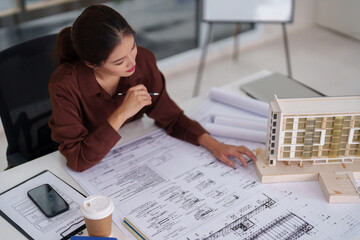 Asian adult woman architect reviews design blueprints building model for new construction project planning professional office workspace