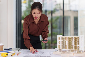 Adult Asian female professional focusing on architectural plans using a smartphone and pen. She meticulously studies design models.