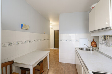 bright kitchen interior featuring white tiled walls, a wooden floor, a simple table and chairs, and a clean sink area. The overall style is functional and minimalist