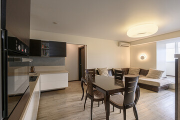 Modern apartment interior. Kitchen with dark cabinets, dining table, and living area with brown sectional. Neutral colors, light wood floors