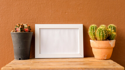 Cacti are placed in pots on a wooden table, with an empty white frame positioned between them. The...