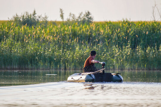 Fisherman driving an inflatable PVC boat with an electric trolling motor on a calm river. Man in an inflatable boat navigating near green reeds bank concept of active weekend, angling and hobby. 