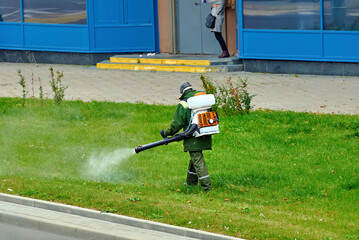 Worker man with backpack mistblower spraying poison insecticide on green grass for tick mosquito and mite control during lawn treatment and outdoor pest prevention maintenance service