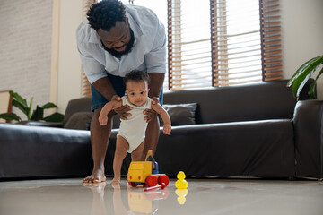 Happy African American dad holding adorable infant son helps him take first steps walking in living room at home.